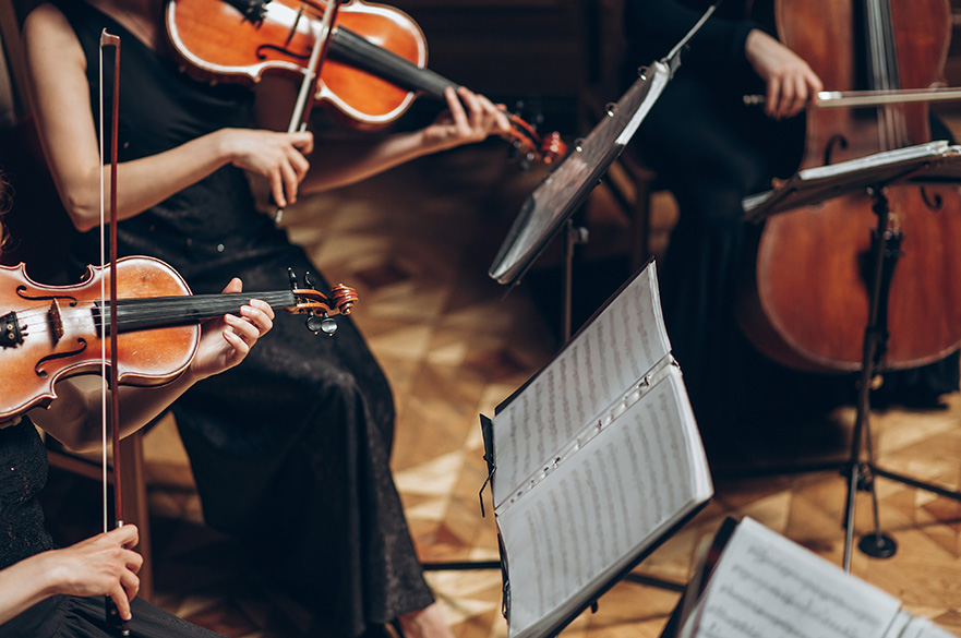 A group of musicians dressed in formal black attire perform seated with violins and a cello, using sheet music stands on a wooden floor.