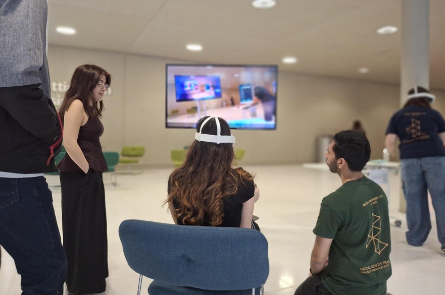 People standing watching while a person sitting tries out VR software. The image they can see in their headset appears on a TV screen.