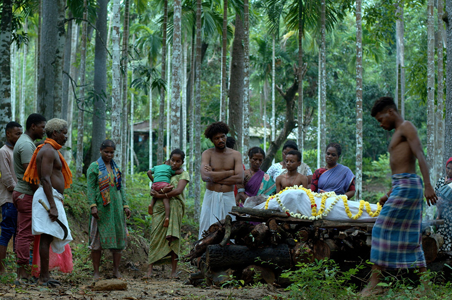 People in traditional attire gathered in a forest around a flower-adorned body on a wooden platform during a ritual