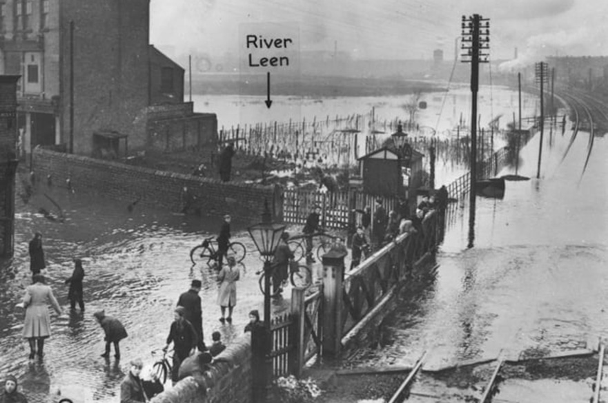 Black and white photograph of a flooded railway crossing with people walking and cycling through the water.