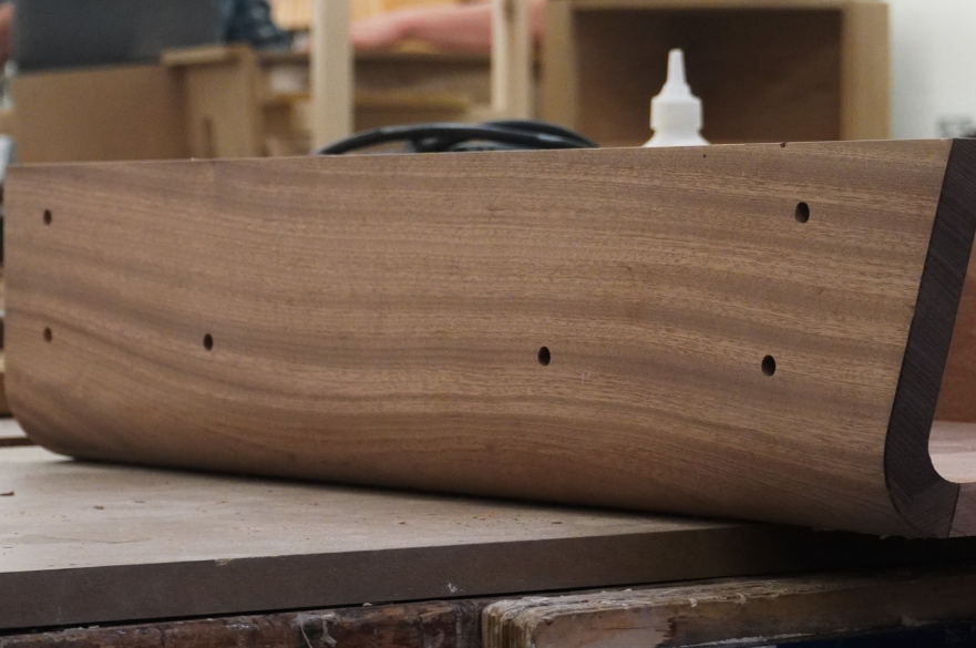 A piece of wood that is being worked on as part of a piece of furniture. Holes have been made in the wood.