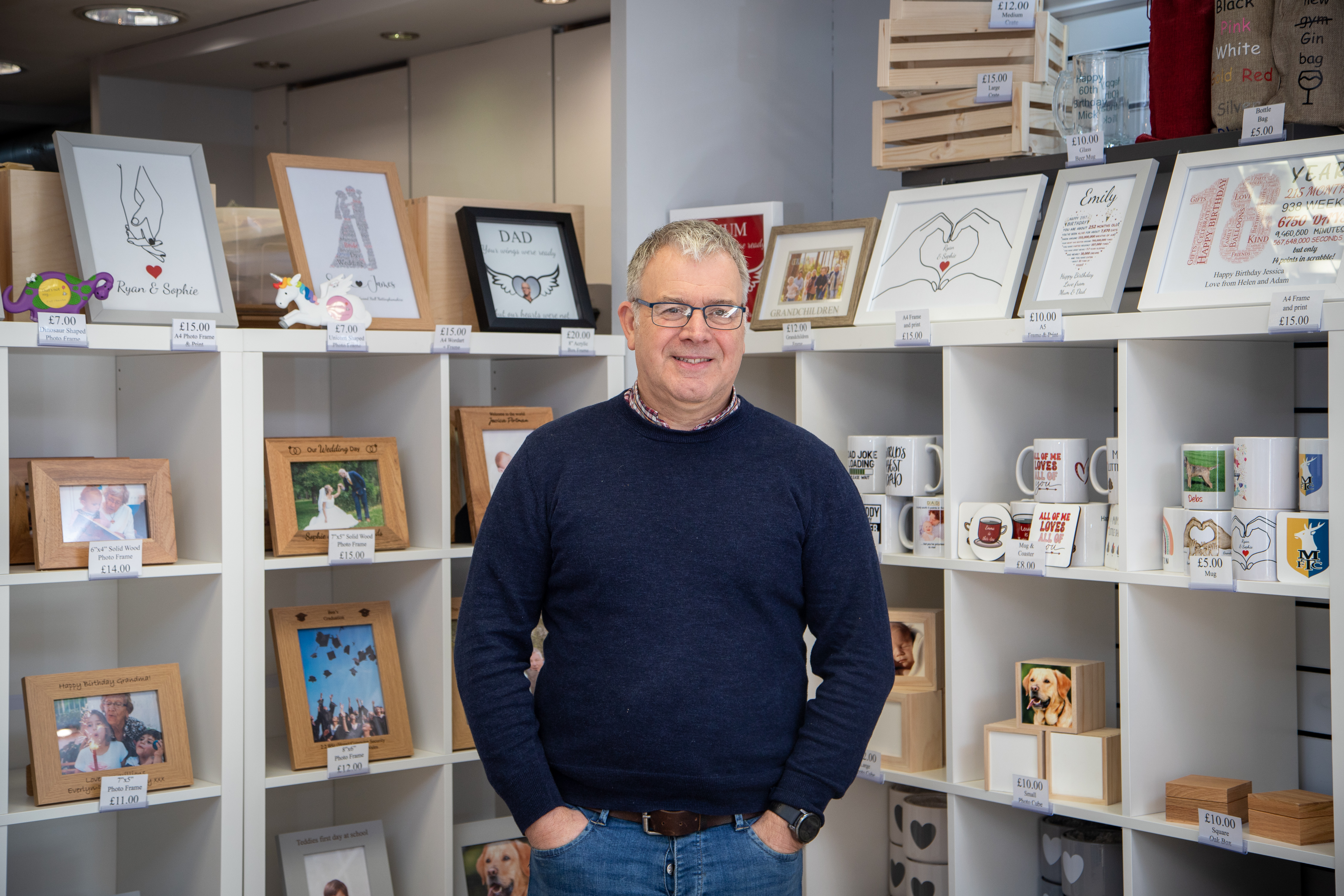 A man in in jeans and a blue jumper stands in front of a promotional display stand full of wooden carved gifts