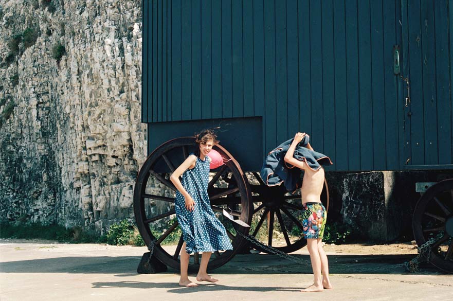 Two children on a beach with a blue wooden cart behind them