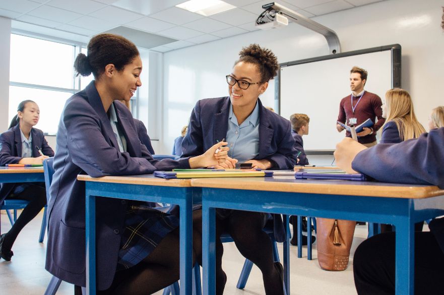 Two school students working together in a lesson while teacher stands at the front of the class