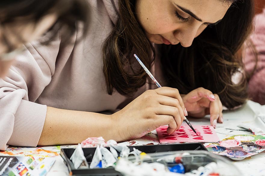 A girl painting a small canvas tile with paints on the table.