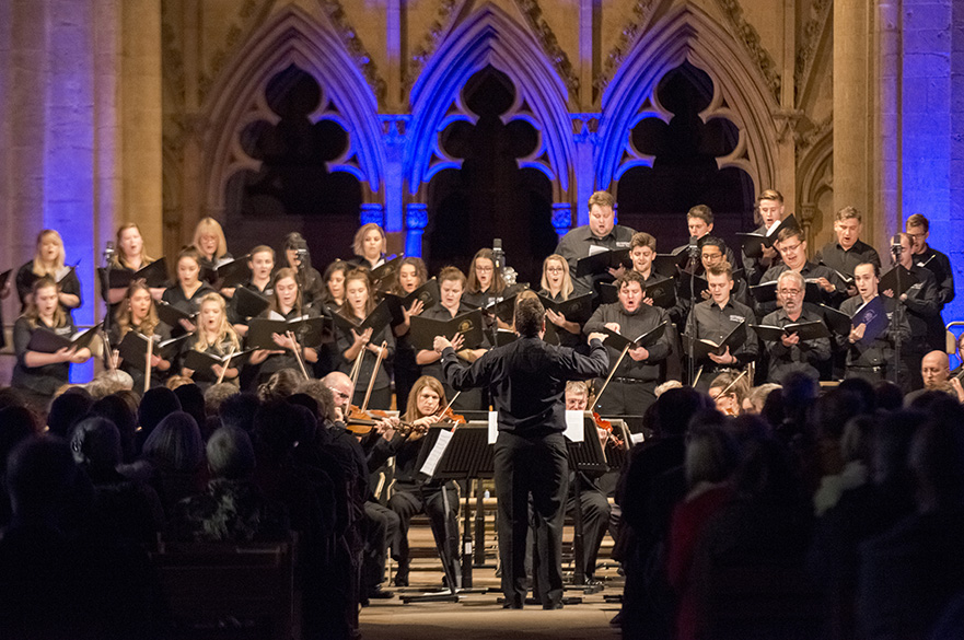 A choir stood singing in a church in front of an ornate stone screen, being conducted by a man.
