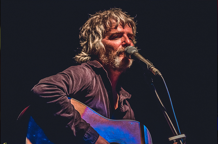 Photograph of John Bramwell singing in a mictrophone playing acoustic guitar.