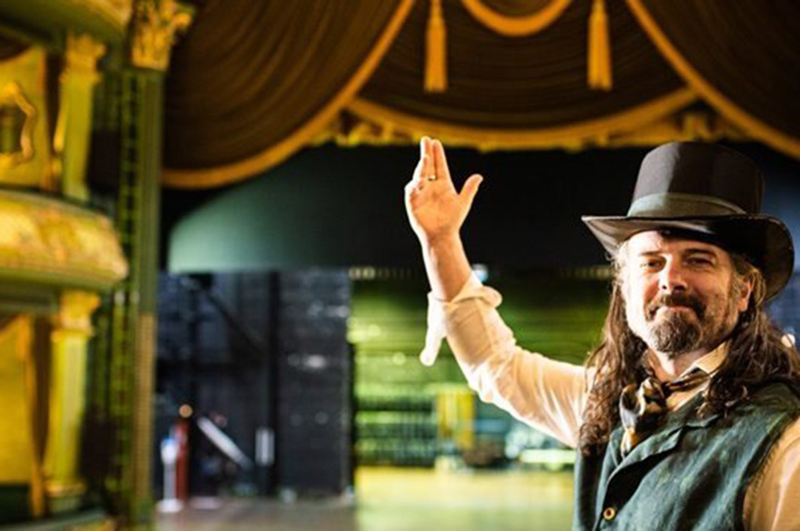 A man wearing a top hat with his arm in the air, stood in front of a theatre stage.