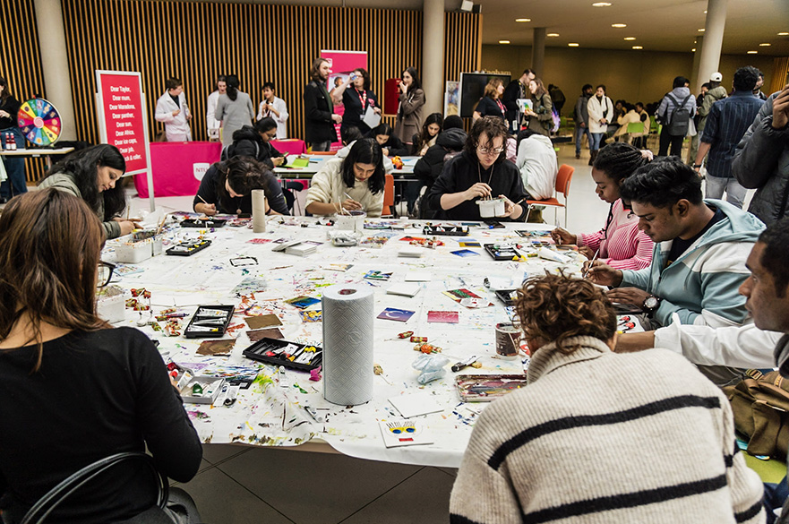 Students sat around a table covered in art supplies, painting colourful tiles.