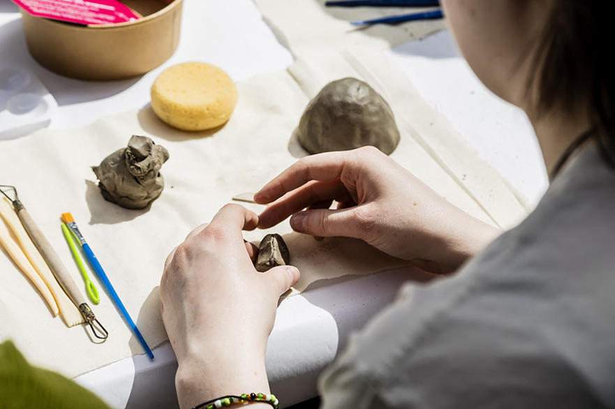 Someone's hands working with a piece of clay on a white cloth. There are small lumps of clay on the table and some modelling tools.
