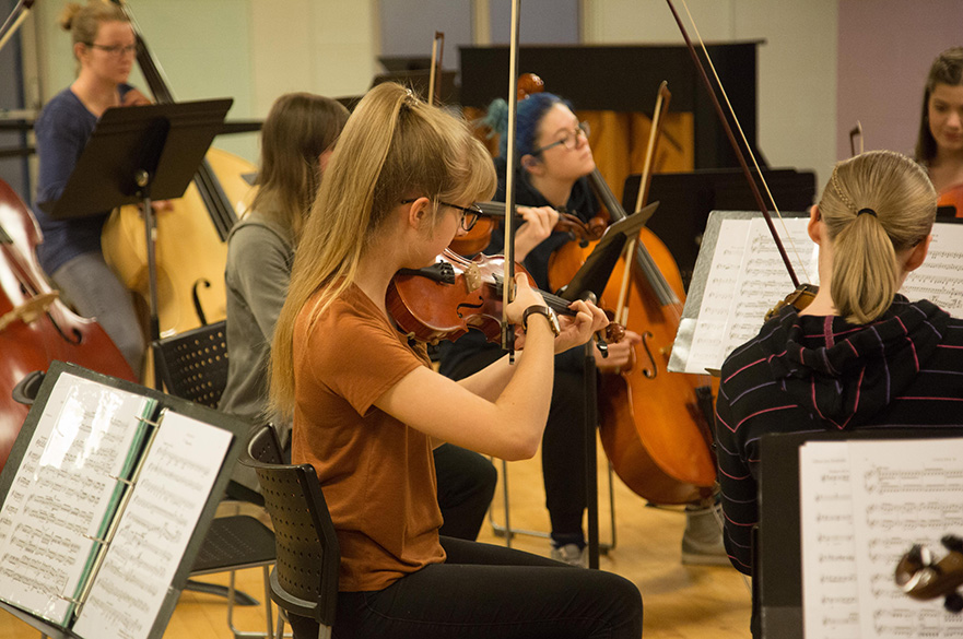 Students playing violins, violas, cellos and double basses in a rehearsal studio.