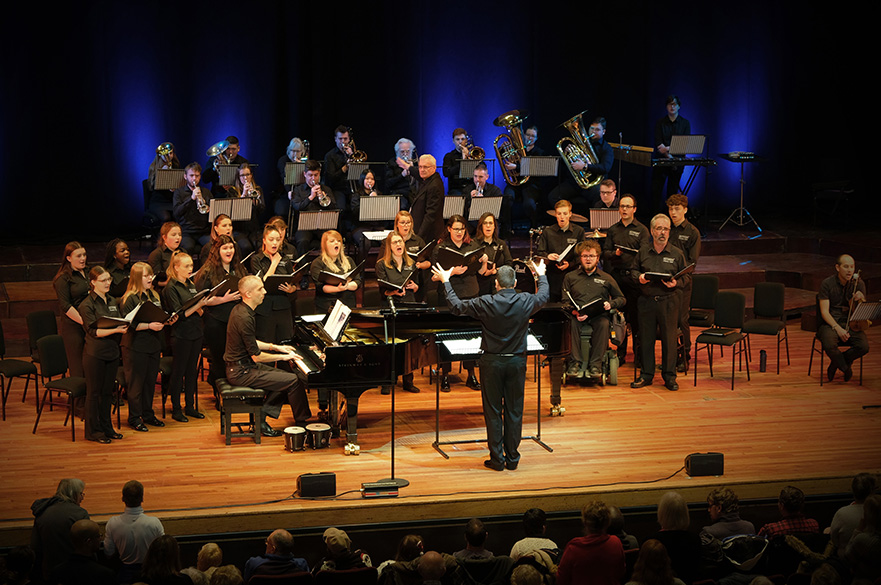 mixed group of singers on stage with a piano and conductor performing at a music concert