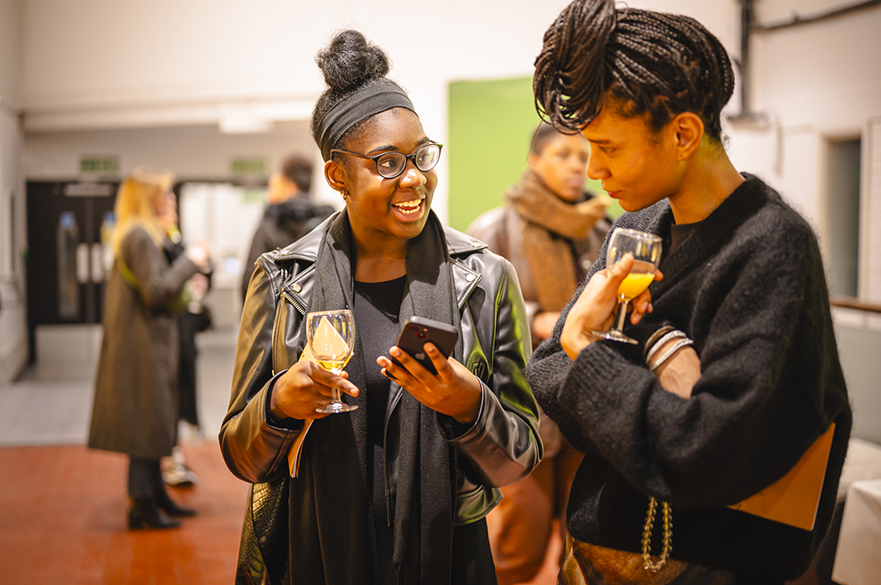 Two girls talking, holding glasses, at an exhibition launch.