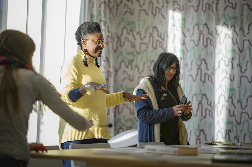 Three women at a gallery exhibition, the curator is a black woman in the middle wearing a yellow sweater. 