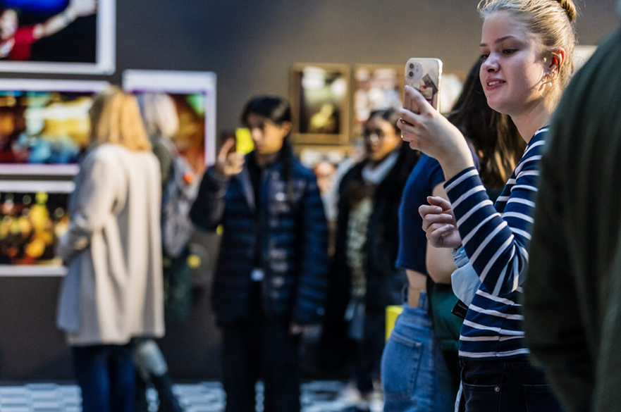 People in an art gallery with a young women near the camera using her phone to photography something out of sight.