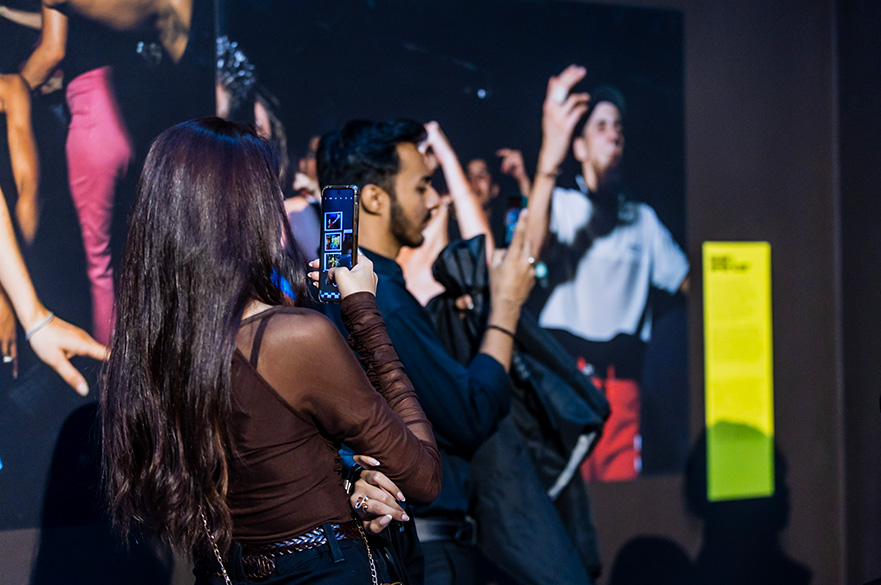 A man and woman taking photos at an art exhibition, with a large image of dancers and an information panel in the background.