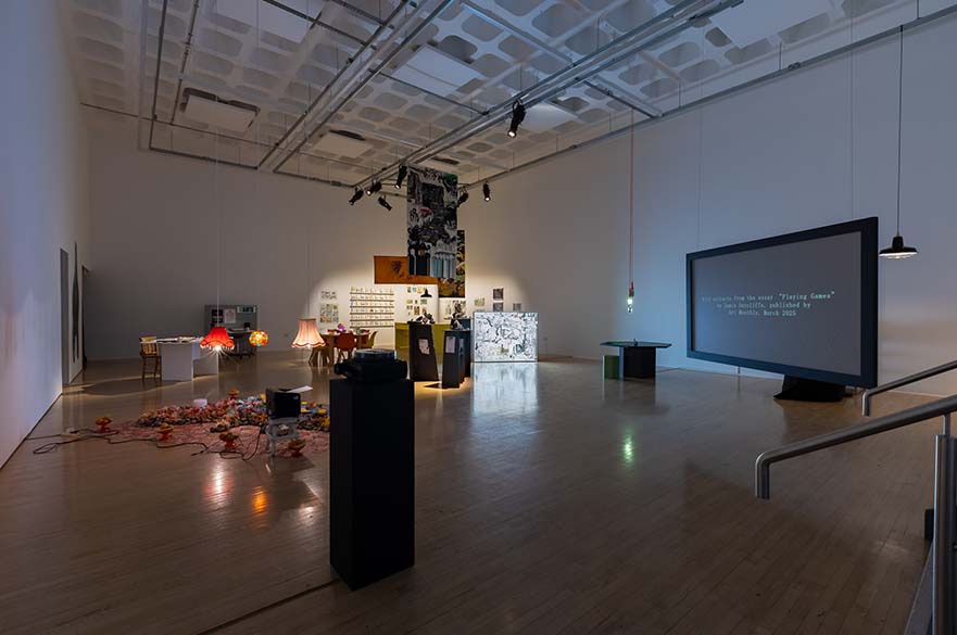 Bonington Gallery's main exhibition space with Weird Hope Engines exhibition on display, including a large screen to the right, hanging banners and lampshades from the ceiling, an artwork on the floor, some desks and artwork on the walls.