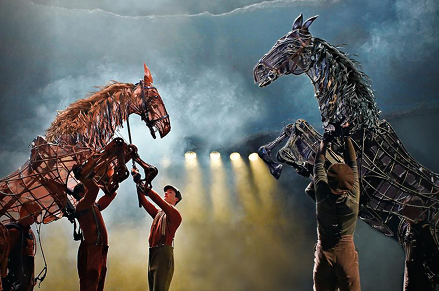Three puppeteers operating two large puppets of horses, on stage.