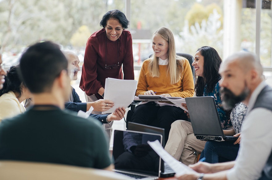 A diverse team looks happy in conversation. They are sitting around with laptops in front of them