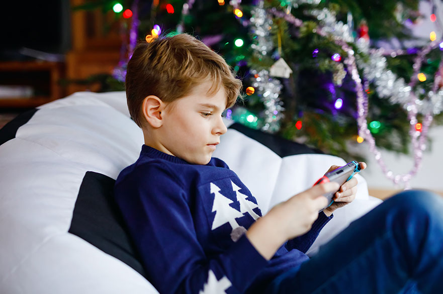 Blond boy playing with a video game on gadget console on Christmas with decorated tree on background
