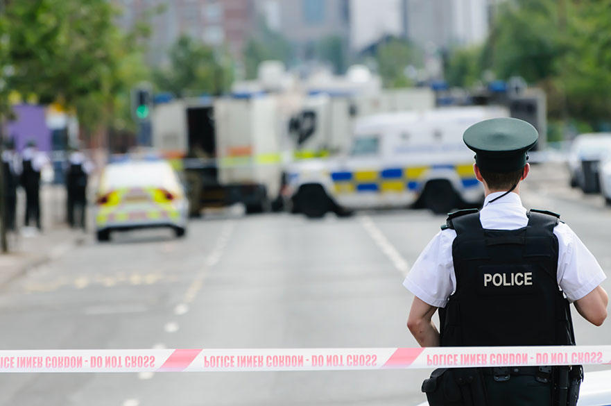 Police officer standing with his back to police tape with police cars in the distance