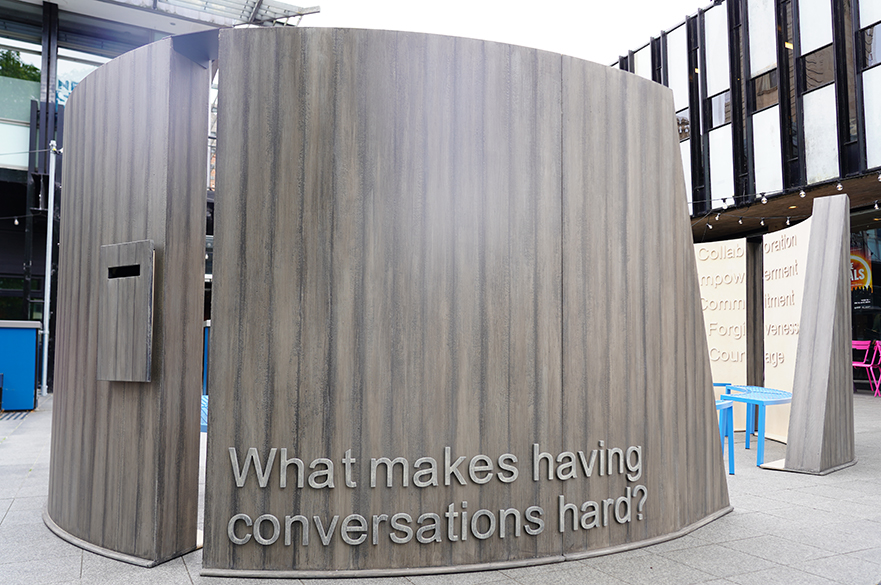 A large circular structure in various pieces, which create a space large enough for a group of people to sit inside, with the words 'What makes having conversations hard?' on the outside.