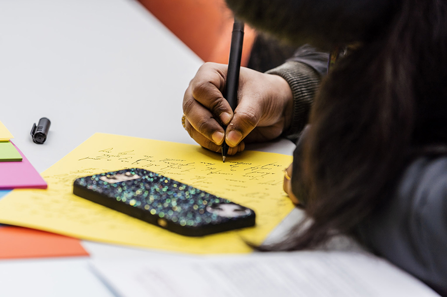 A close up photo of a person writing on a bright yellow sheet of A4 paper with a pen, with their smartphone (with a glittery cover) face down on the table.