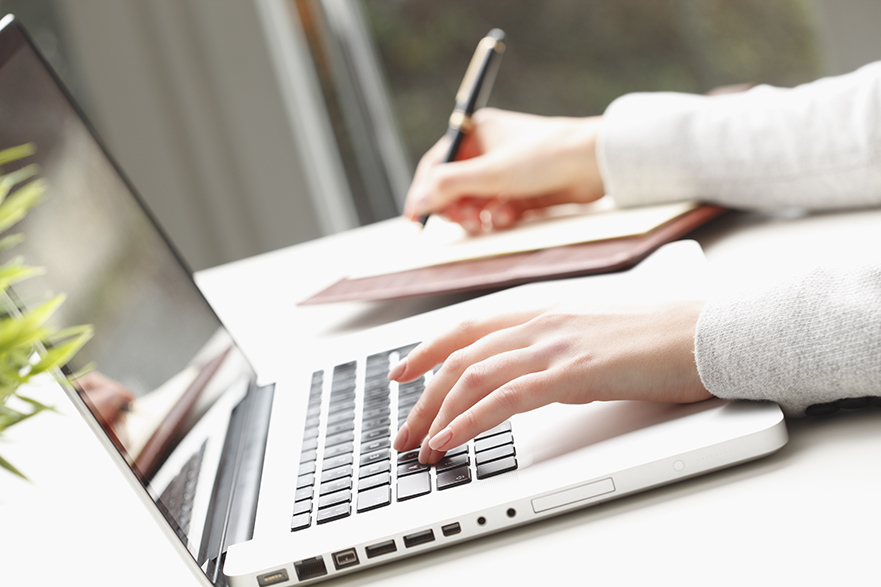 A hand is typing on a laptop whilst the other hand is writing in a notebook. 