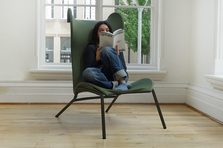 Green and wooden lounge chair displayed next to a tall window with the sun shining through and a woman sitting on it reading a book