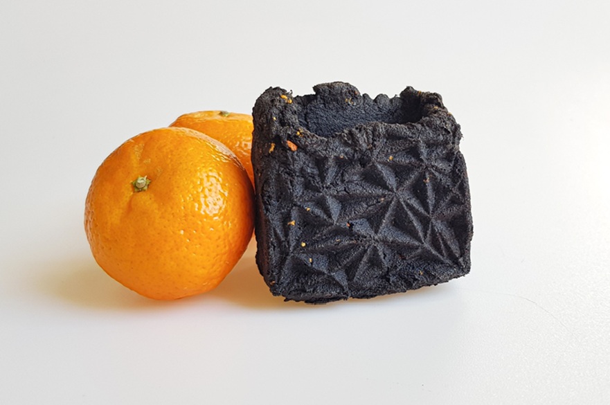 An orange-like fruit and next to it a container that has been made out of dried fruit and coffee grounds. They are on a plain white table.