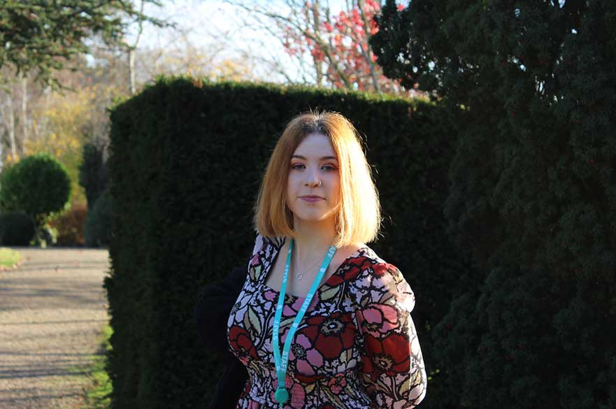 Lorna Shorten-Paice stands in the sun outdoors, with some dark green bushes behind her.