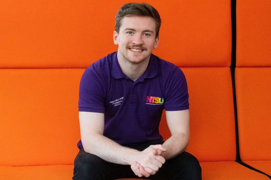 Benedict Wills, wearing an NTSU polo shirt, sitting on a bright orange sofa,
