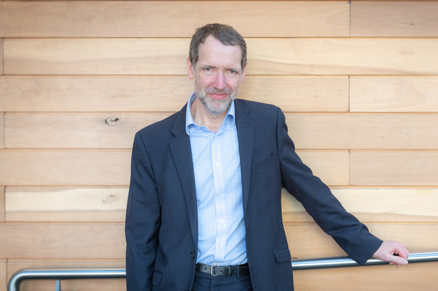 The image shows governor Jon Scott in a jacket and an open-neck shirt, his left hand resting casually on a railing.
