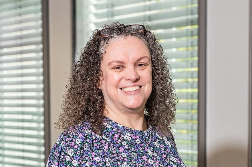 Sara Murcott stands smiling in front of a window with blinds across it.