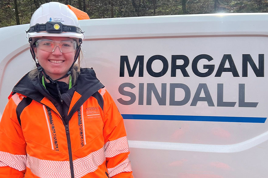 Rebecca wearing her construction gear standing in front of a Morgan Sindall sign